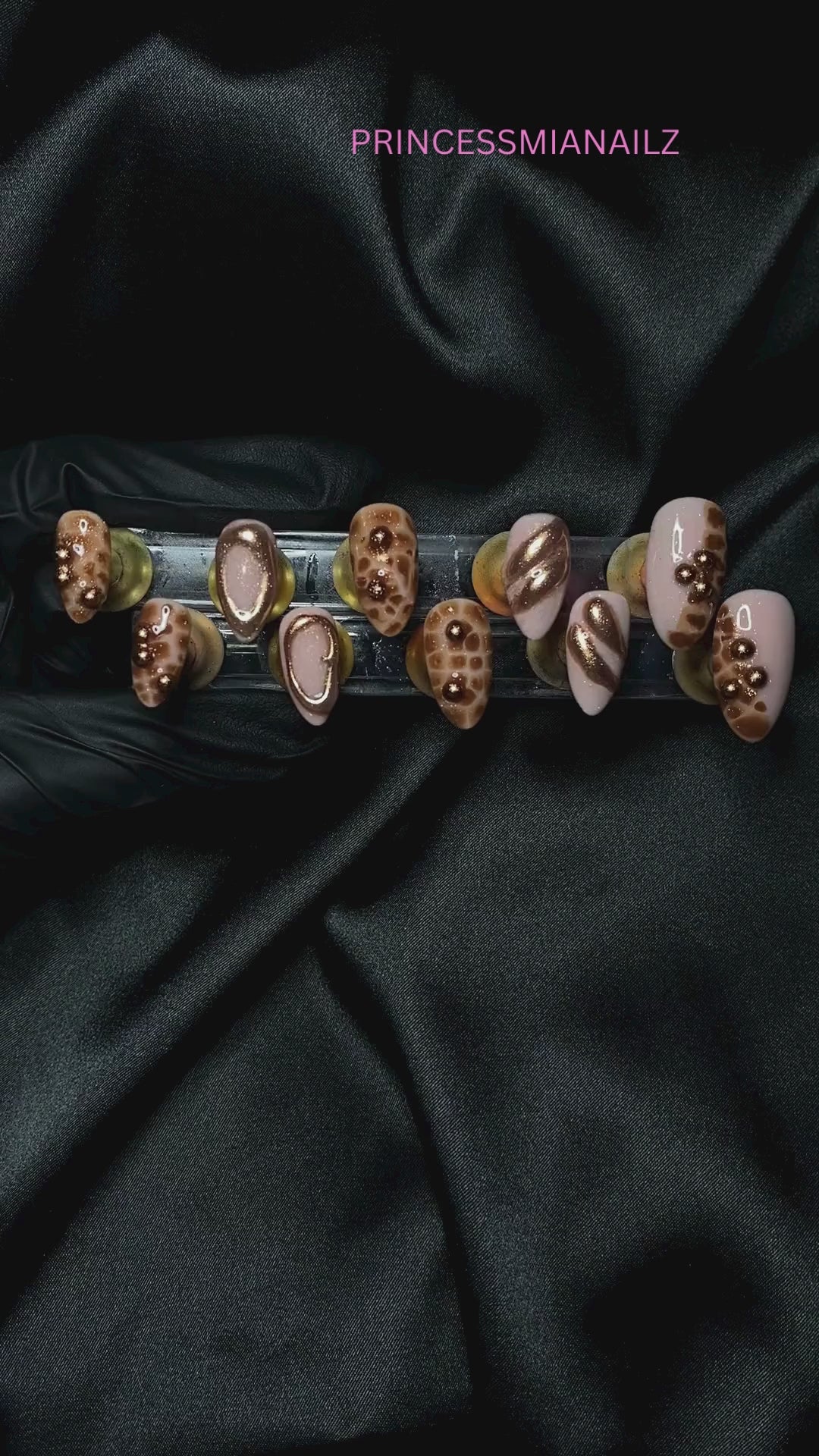 Cinnamon-hued press-on nails by PrincessMiaNailz, displayed on a luxurious black fabric for a regal aesthetic.